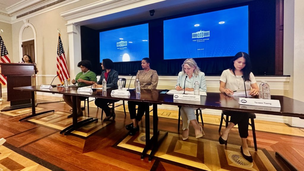 Five individuals in business attire sitting at a panelist table with two tv screens behind them that have a blue graphic with a white white house logo. On individual stands at the podium flanked with two USA flags in the left side of the photo