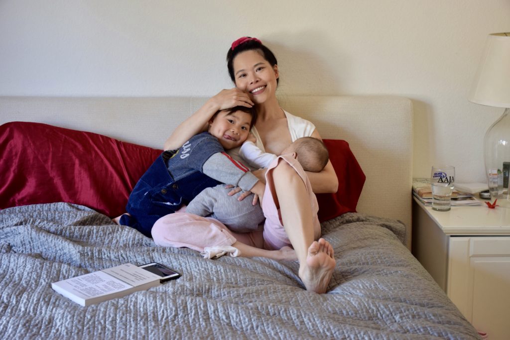 Photo of Chinese woman sitting on a bed with a small child laying on her left while breastfeeding an infant cradled in her left arm