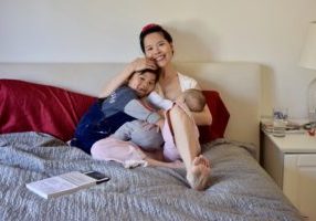 Photo of Chinese woman sitting on a bed with a small child laying on her left while breastfeeding an infant cradled in her left arm